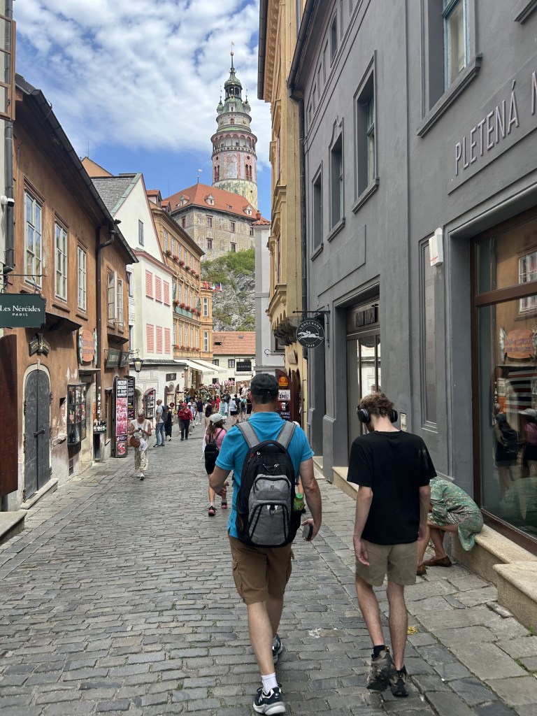 A cobblestone street lined with shops and people walking, featuring a historic tower in the background against a partly cloudy sky.
